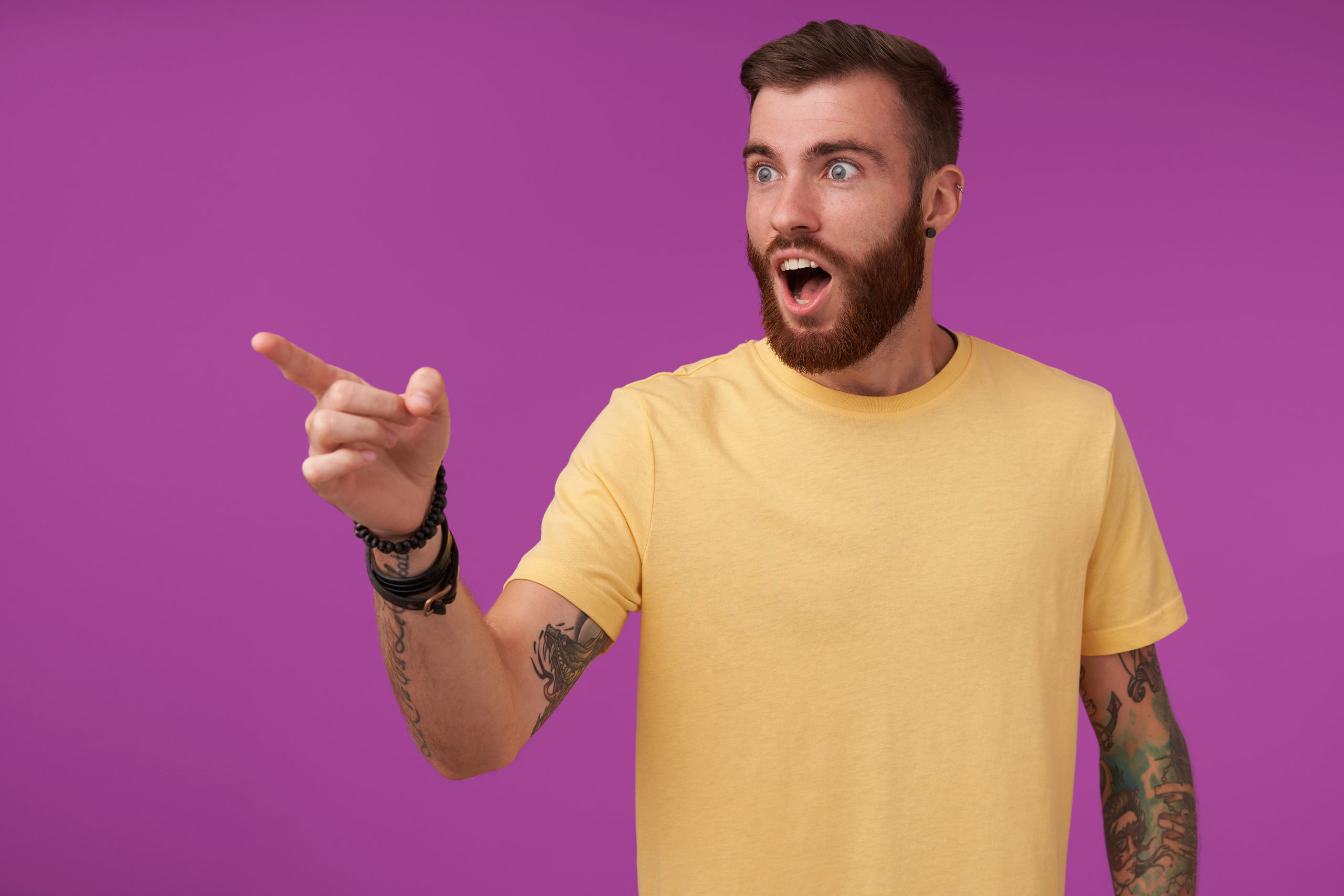 Shocked handsome brunette bearded guy with tattooes wearing yellow t-shirt and fitness watch noticing something and showing on it with forefinger, isolated over purple background